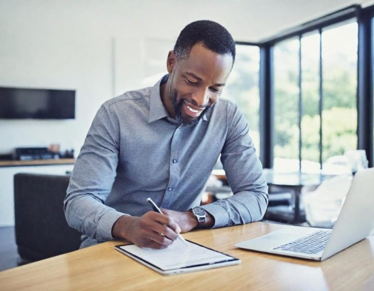 Smiling man writing notes at a table with a laptop nearby and a window view.